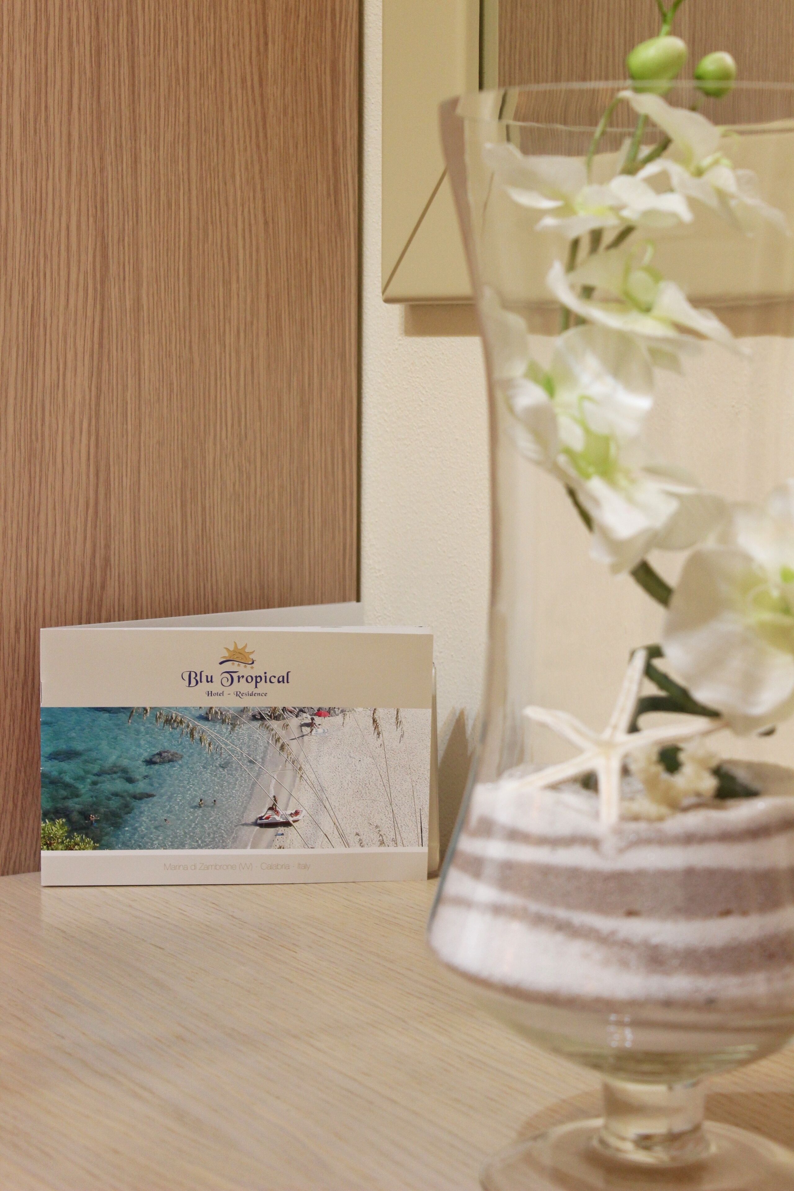 Brochure di un hotel, 'Blu Tropical Hotel - Residence', con una veduta costiera e un bicchiere di dessert decorato con fiori bianchi.