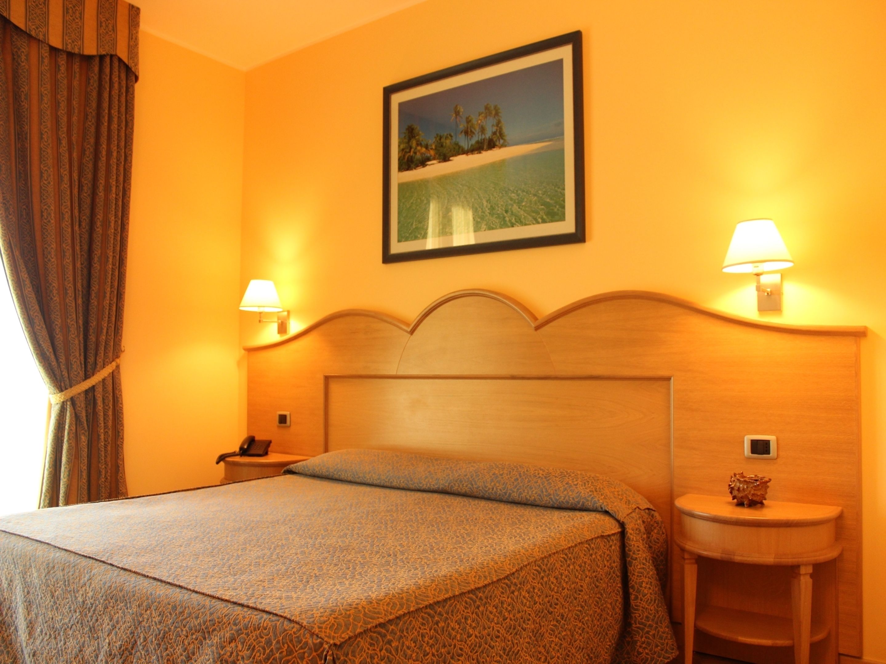 Quarto de hotel com uma cama, duas luminárias de cabeceira, uma mesa de cabeceira, um quadro na parede e uma janela com cortinas.