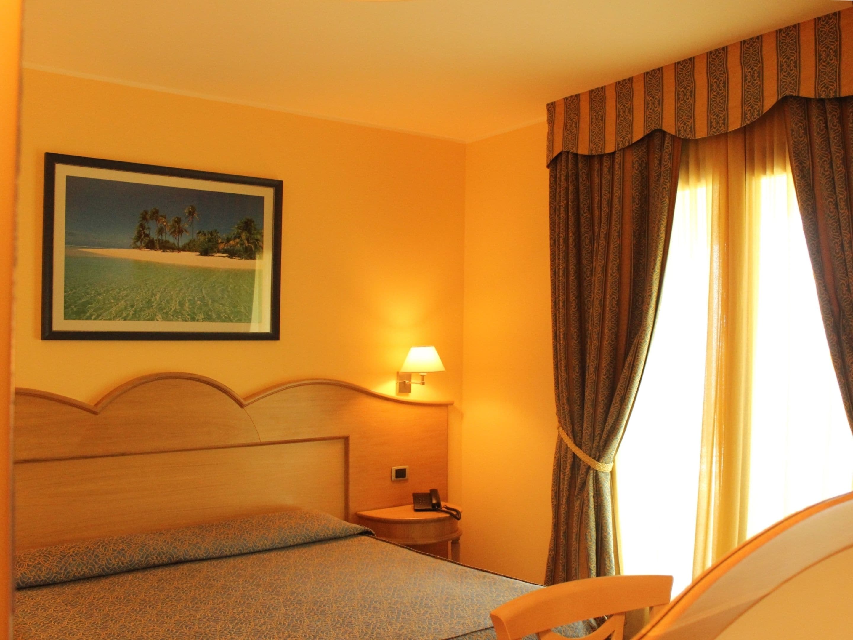 A hotel room with a bed, a nightstand, a lamp, a framed picture on the wall, and a window with curtains.