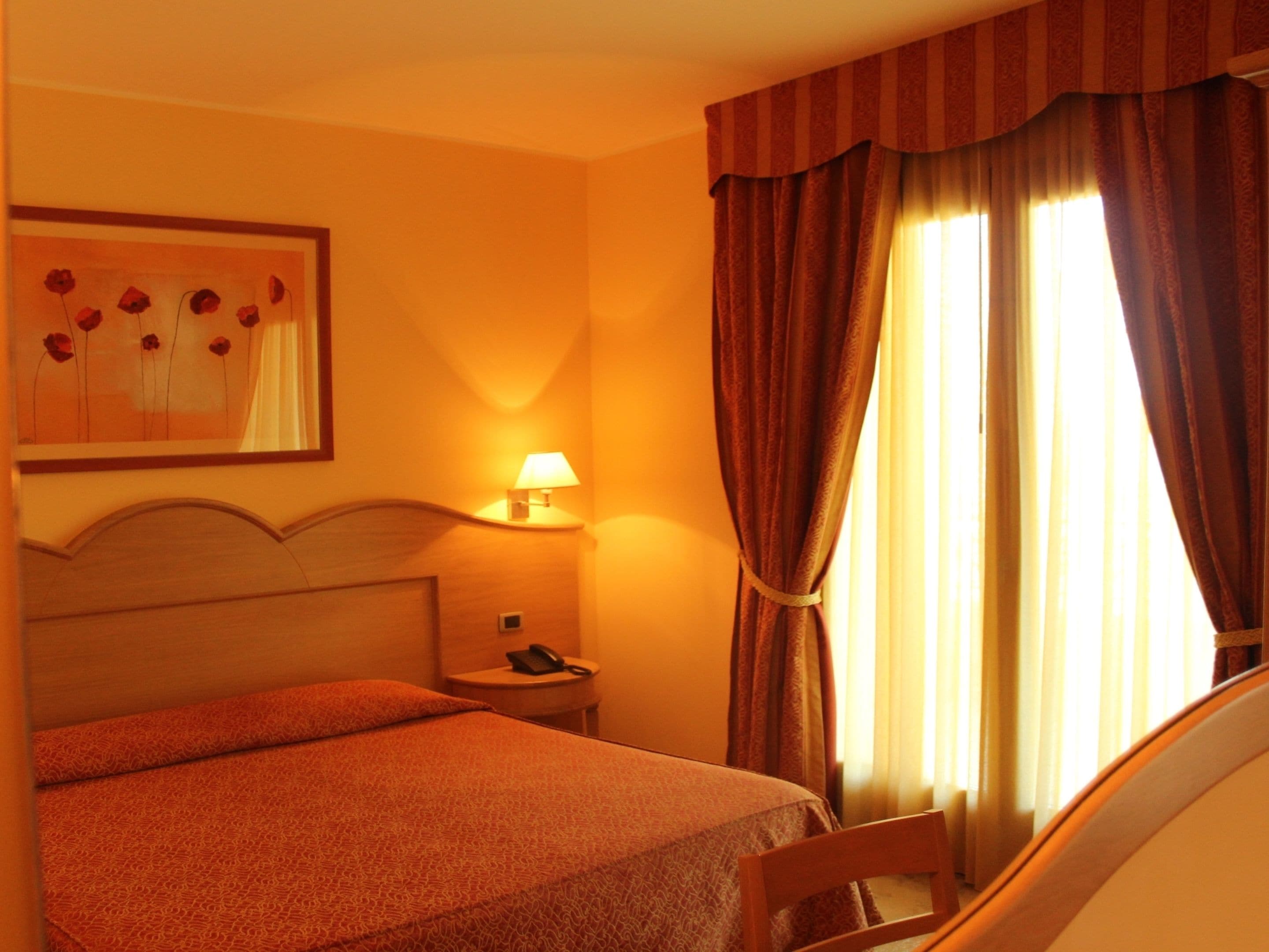 A hotel room with a bed, a nightstand, a lamp, a painting on the wall, and curtains on the window.