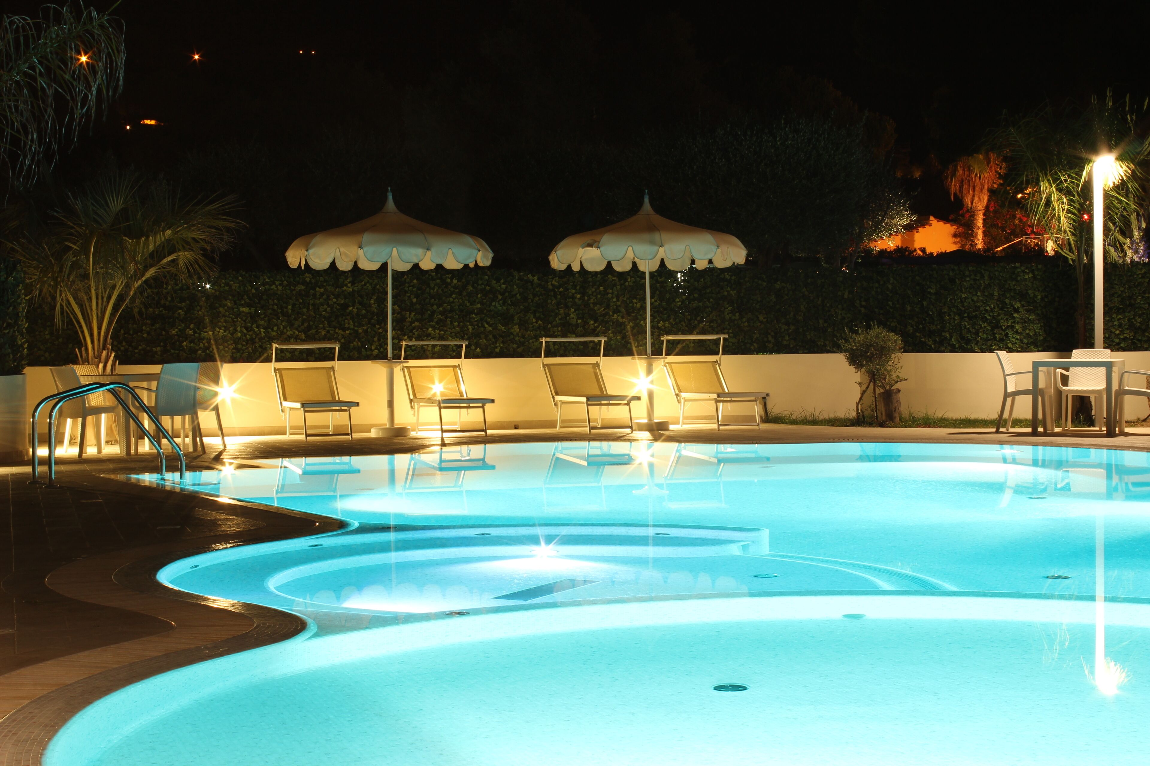 Una piscina nocturna con sillones de descanso y sombrillas, rodeada por un seto y un muro.