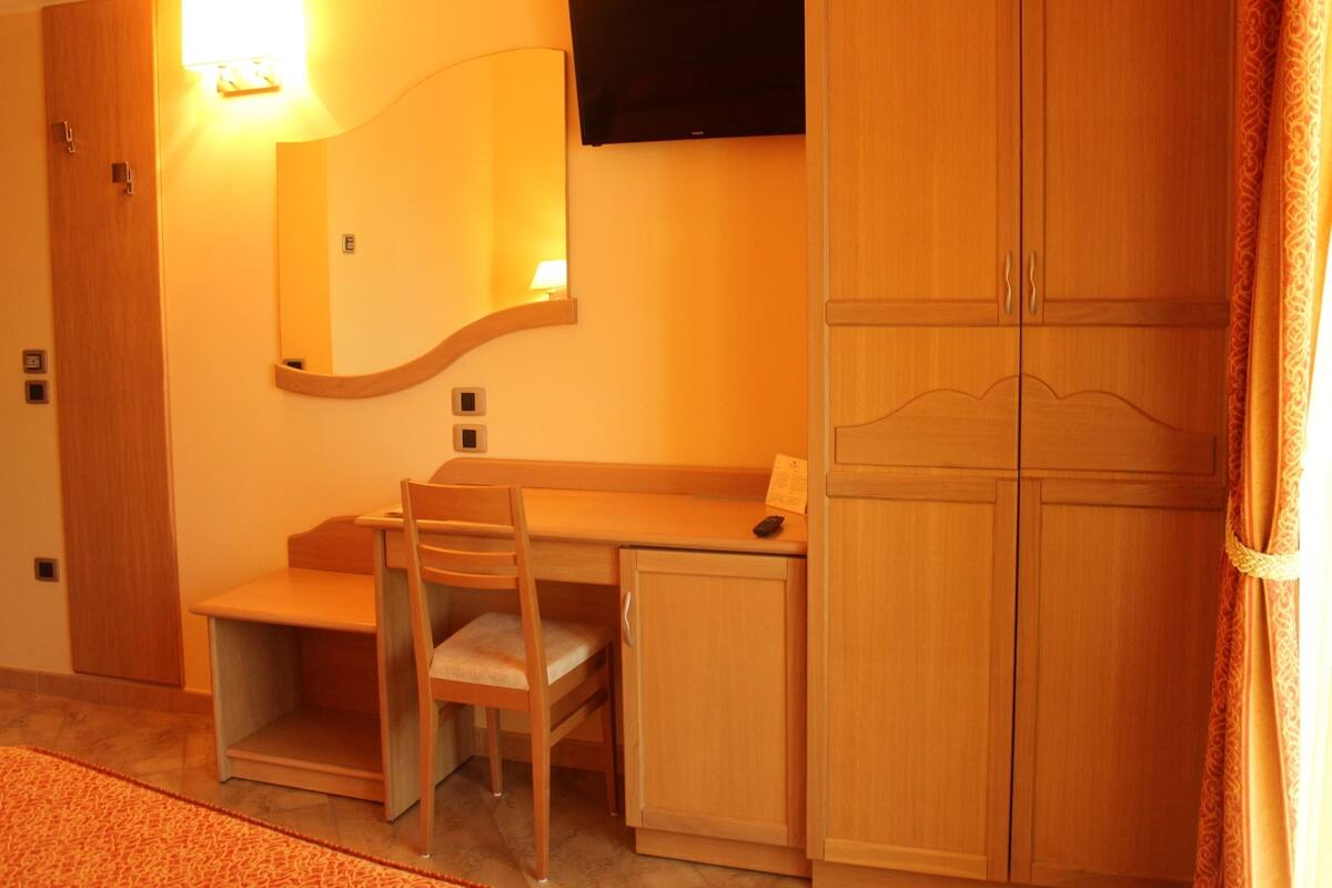 A hotel room with a wooden desk, a chair, a television mounted on the wall, and a wardrobe.