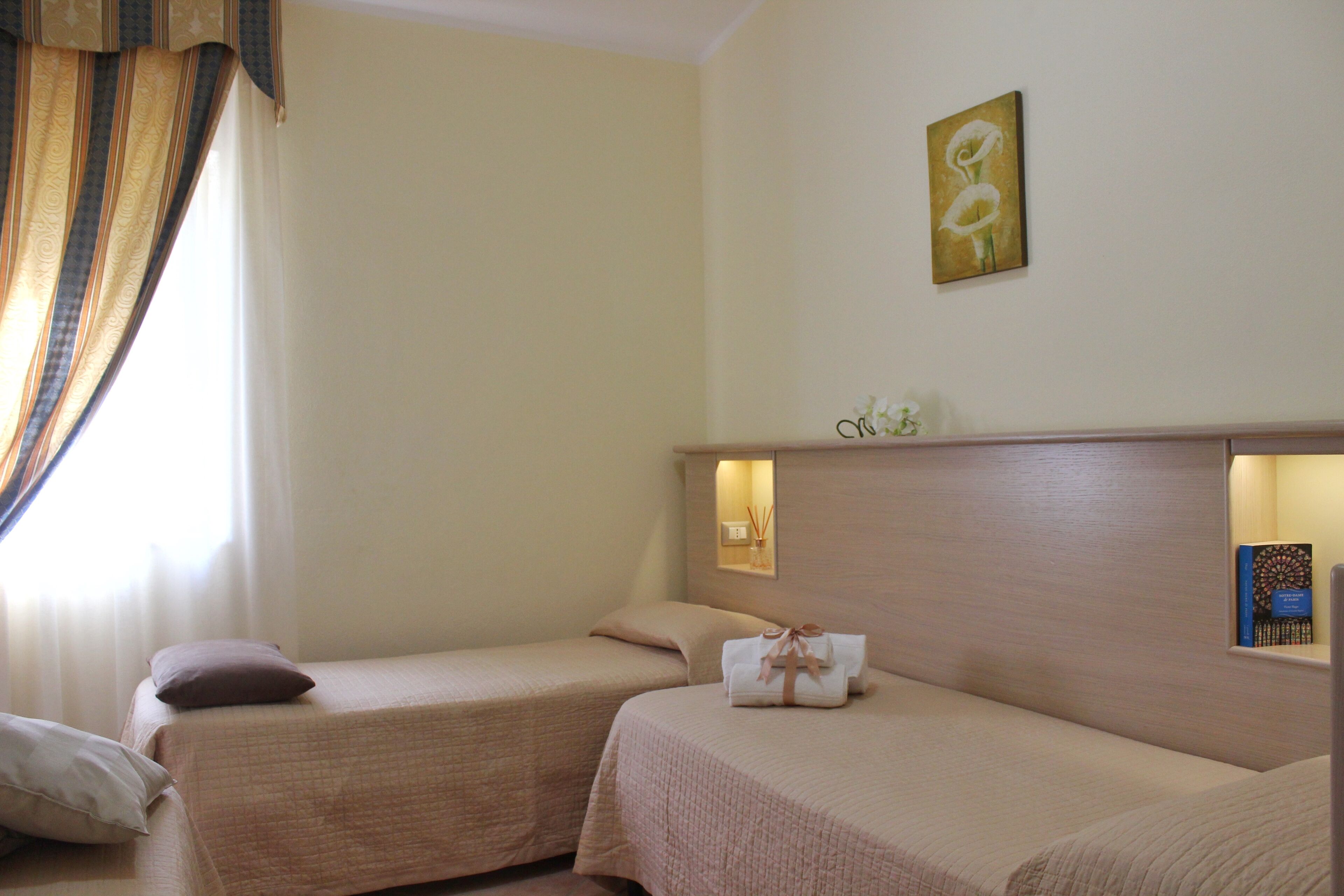 A hotel room with two single beds, a bedside table with a lamp, a wall-mounted artwork, and a window with curtains.