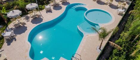 A large, uniquely shaped swimming pool surrounded by lounge chairs and umbrellas, with a well-maintained garden and a pergola in the background.