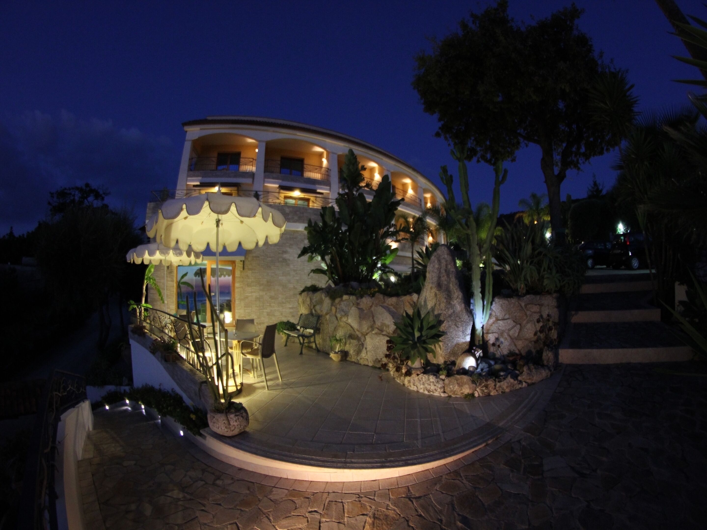 Un edificio grande y curvo con varios balcones, un sendero de piedra que conduce a la entrada y jardines tropicales.