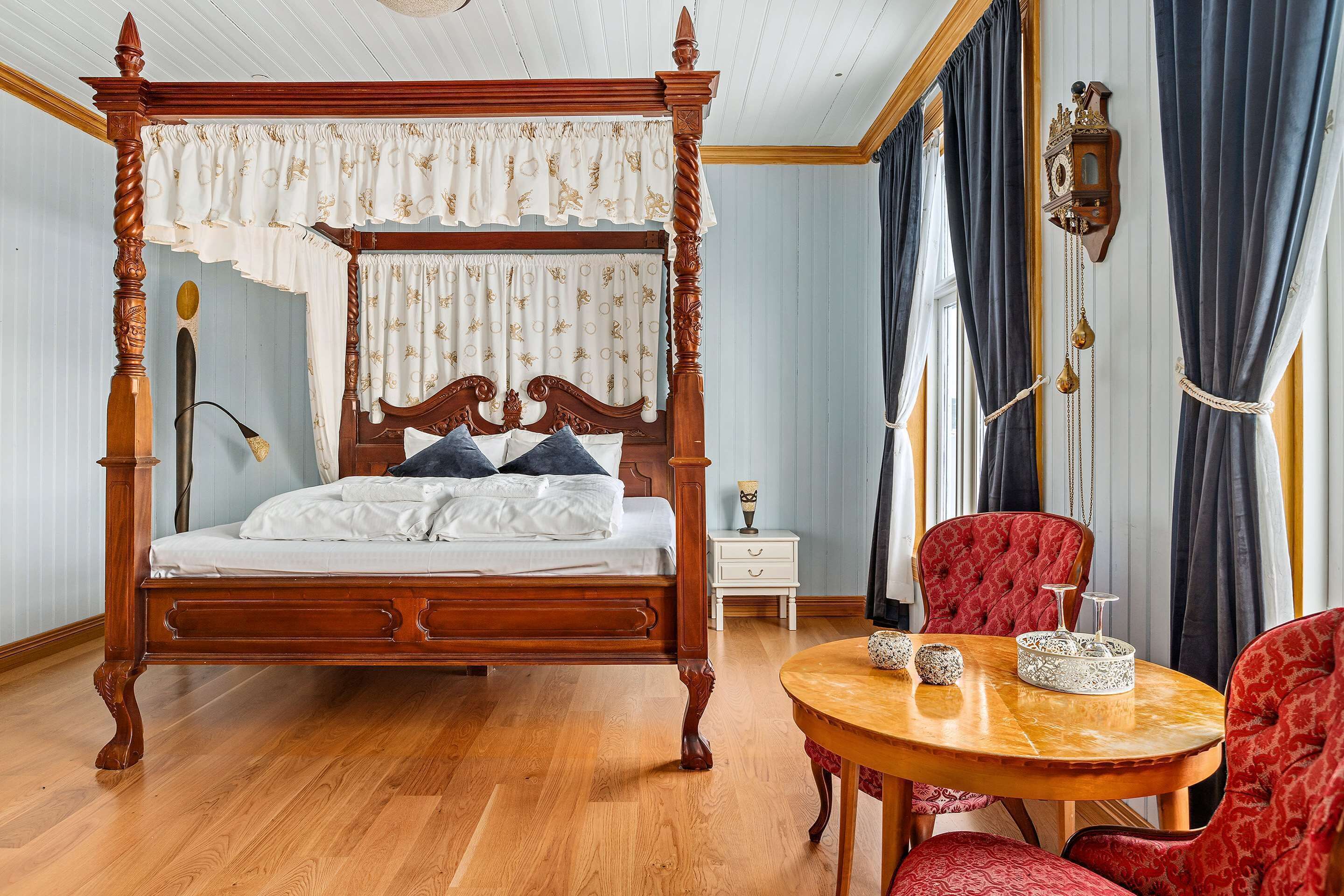 A four-poster bed with a canopy, a round wooden table with chairs, and a nightstand with a lamp.