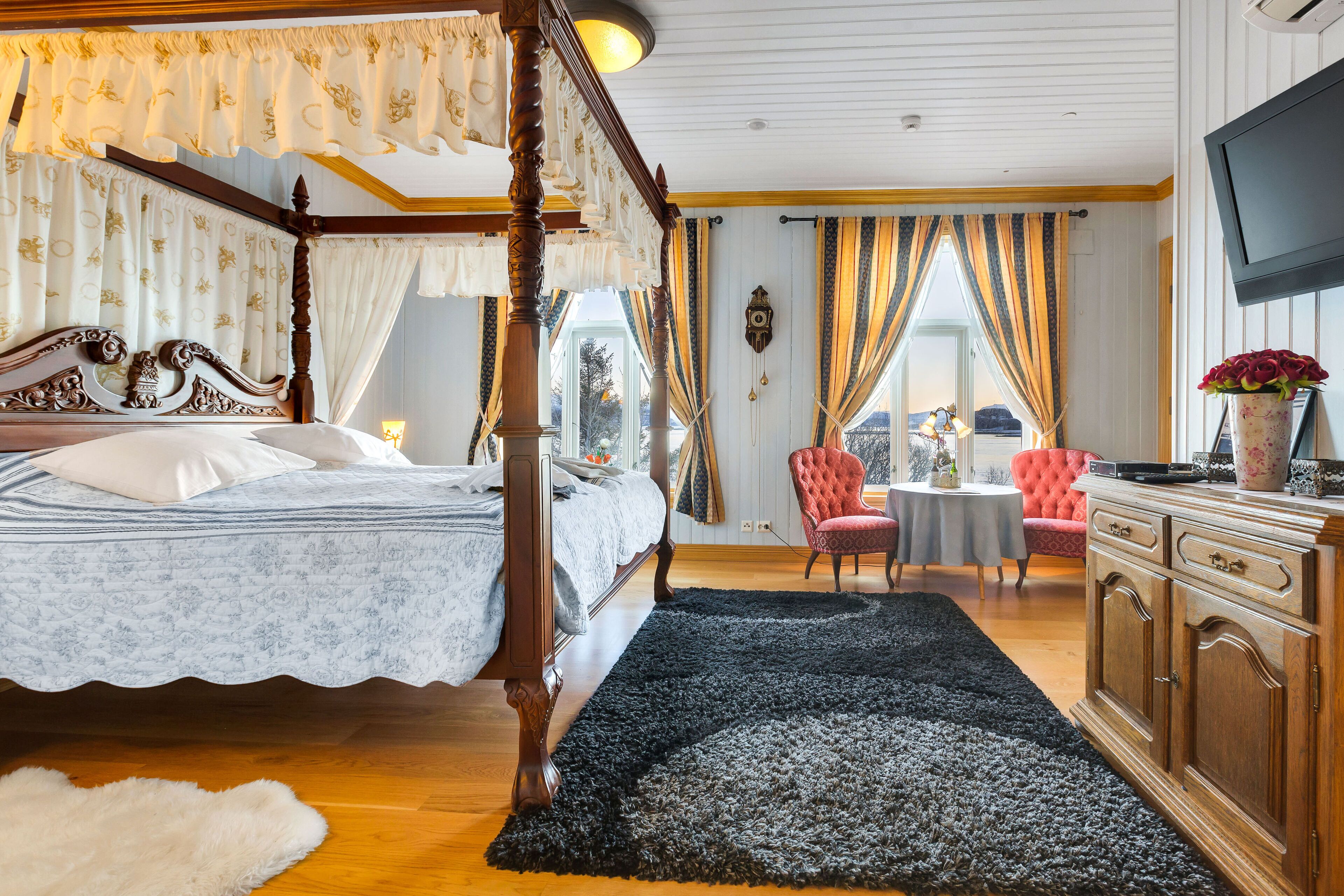 Un lit à baldaquin avec une couvre-lit à motifs blancs et bleus, un tapis bleu foncé, une commode en bois, un téléviseur fixé au mur et un fauteuil rouge dans une pièce dotée de grandes fenêtres et de rideaux.