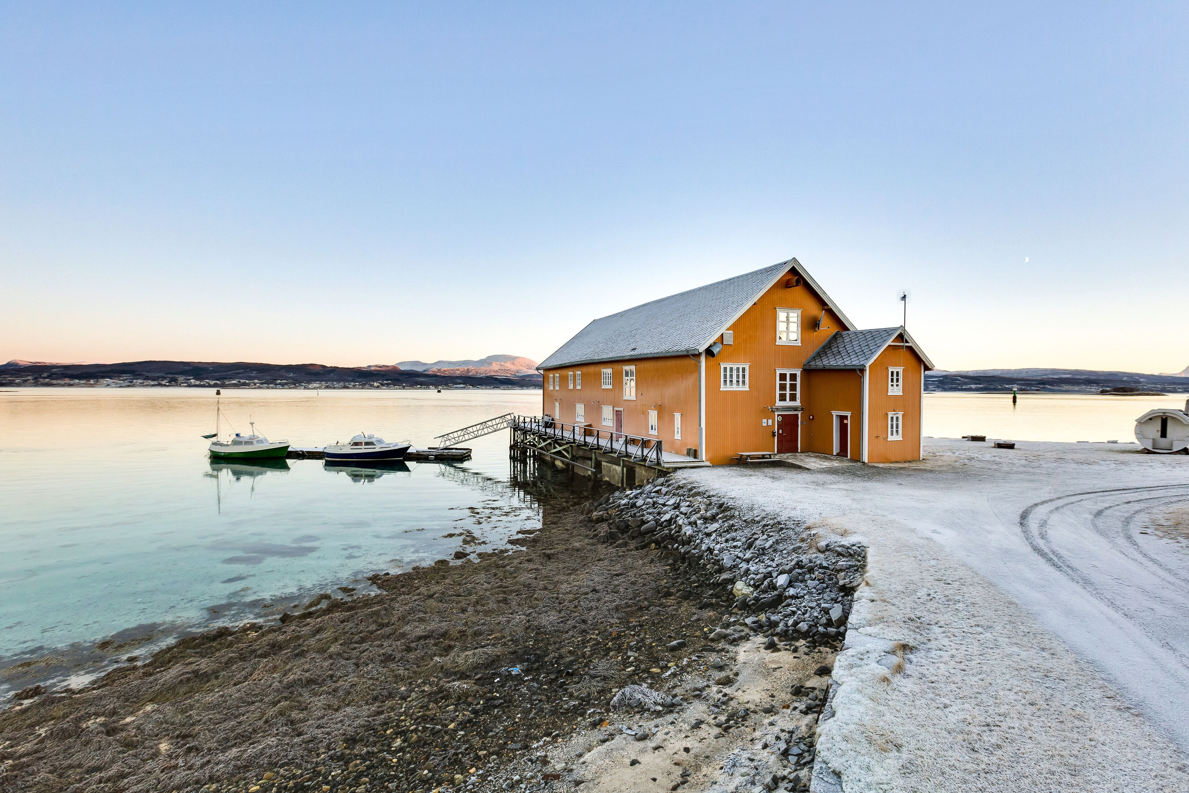 Un bâtiment situé sur la côte, doté d’un quai, de bateaux et offrant une vue dégagée sur l’eau et les montagnes lointaines.
