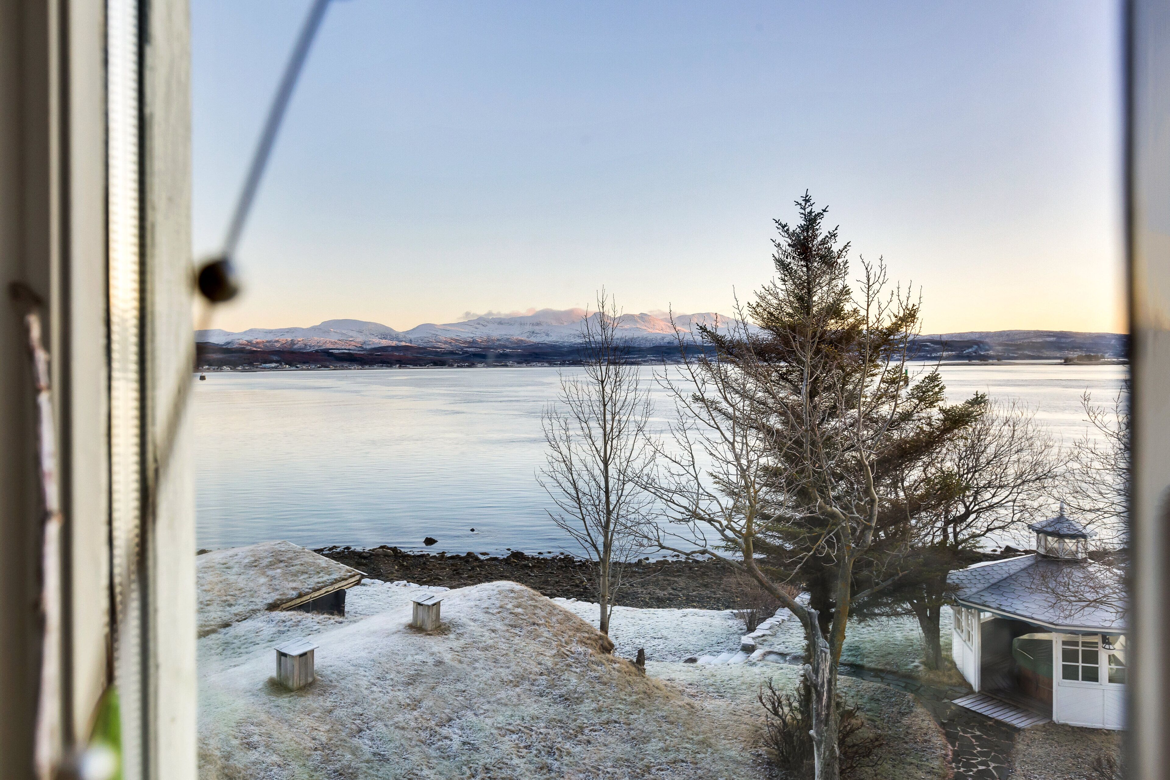 A view of a lake with mountains in the background, a snow-covered ground, and a small building near the shore.