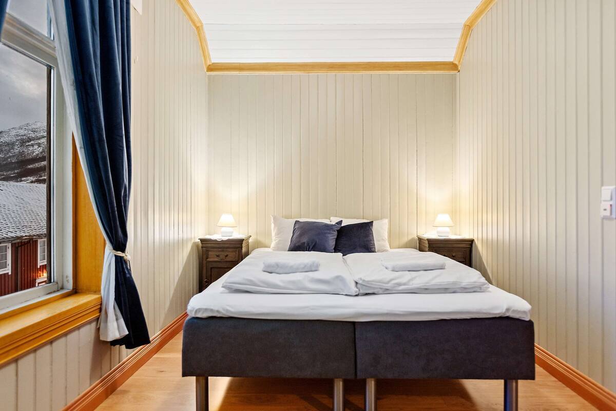 Siististi pedattu sänky valkoisilla lakanoiden ja tyynyjen kanssa, kaksi yöpöytää lamppuineen, ikkuna verhoineen ja näkymä ulkona olevaan rakennukseen.