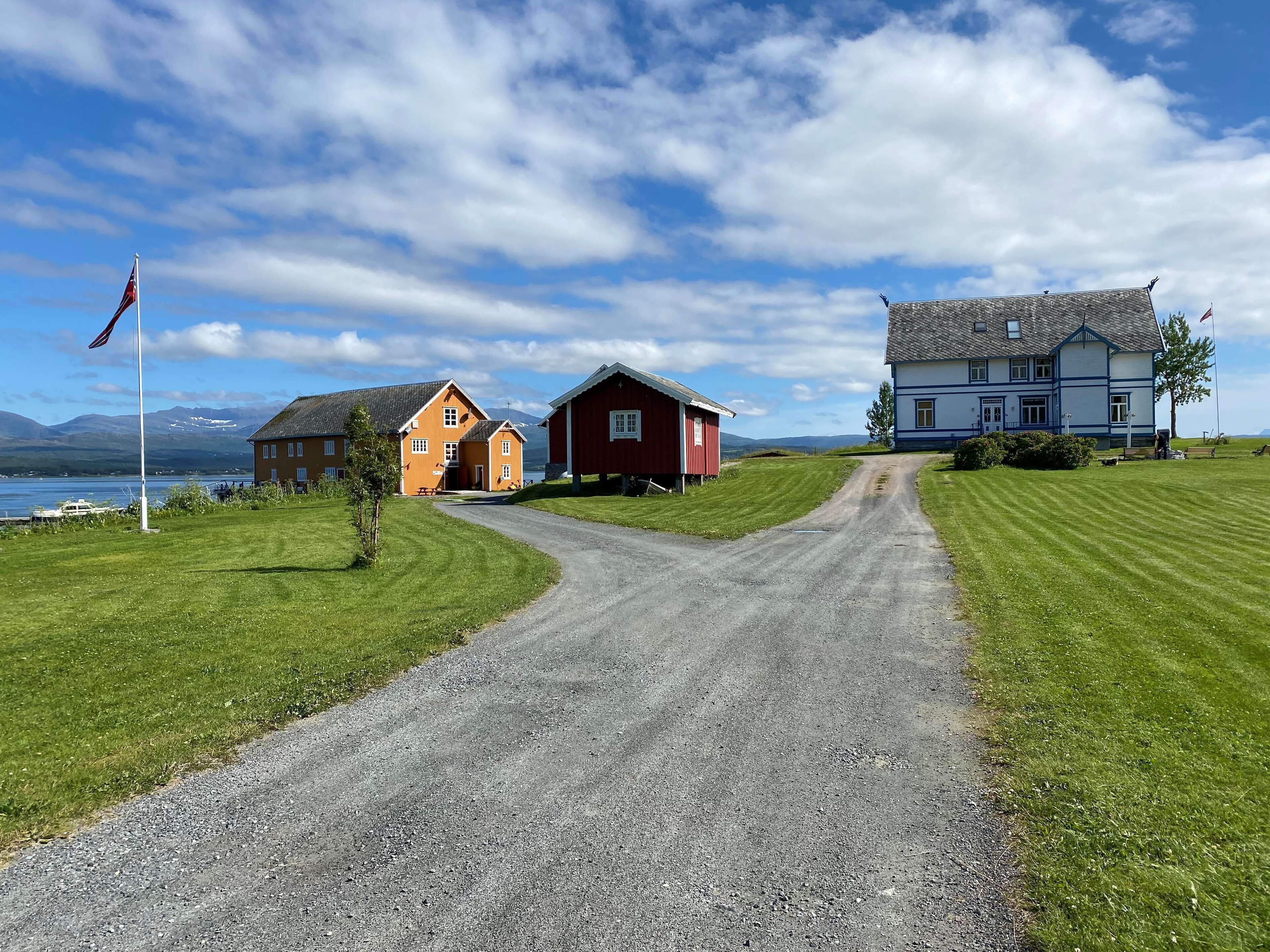 A gravel road leading to a row of houses with a flagpole, a body of water, and mountains in the background.