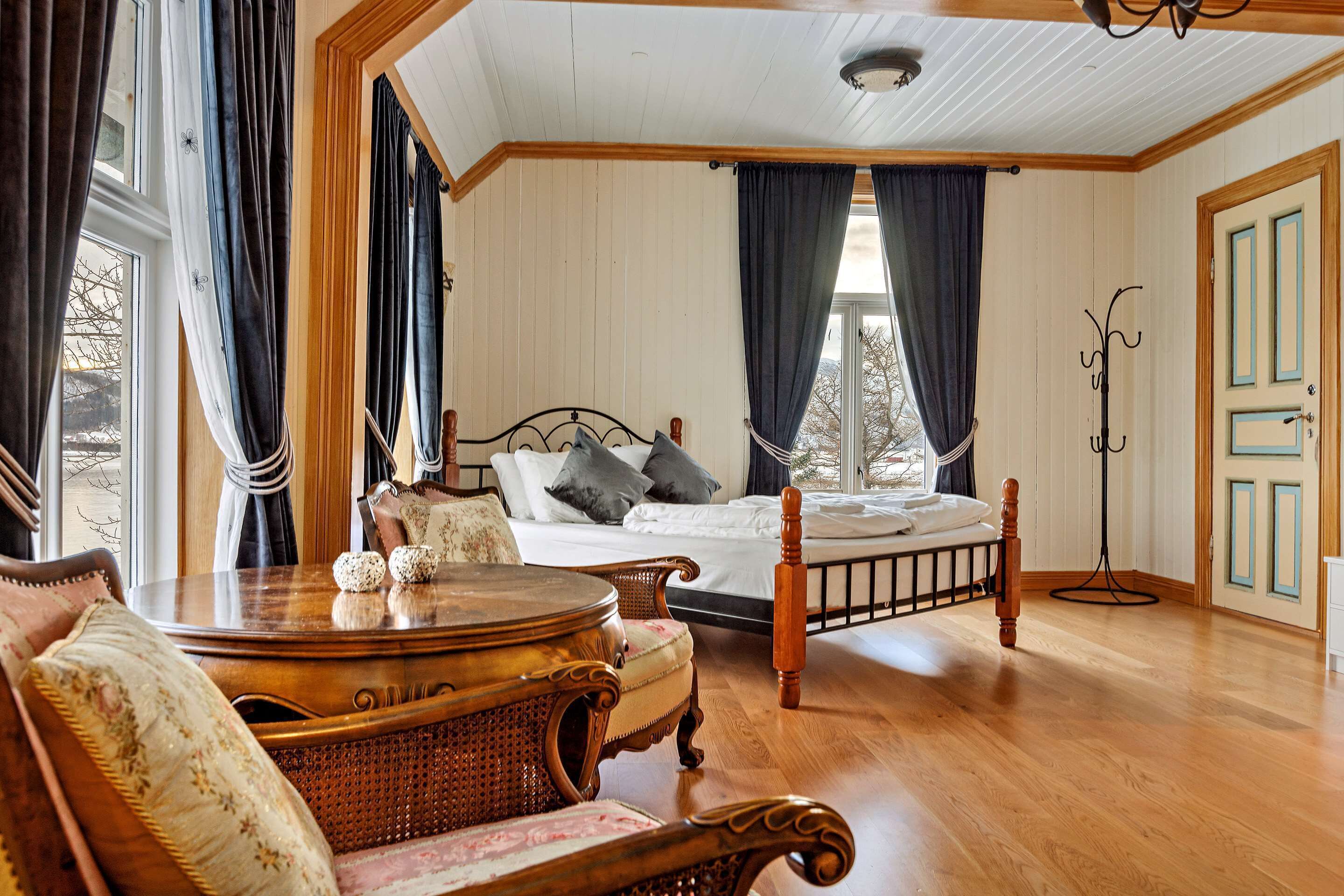 A bedroom with a wooden floor, a four-poster bed, a bedside table, a chair, and a floor lamp.