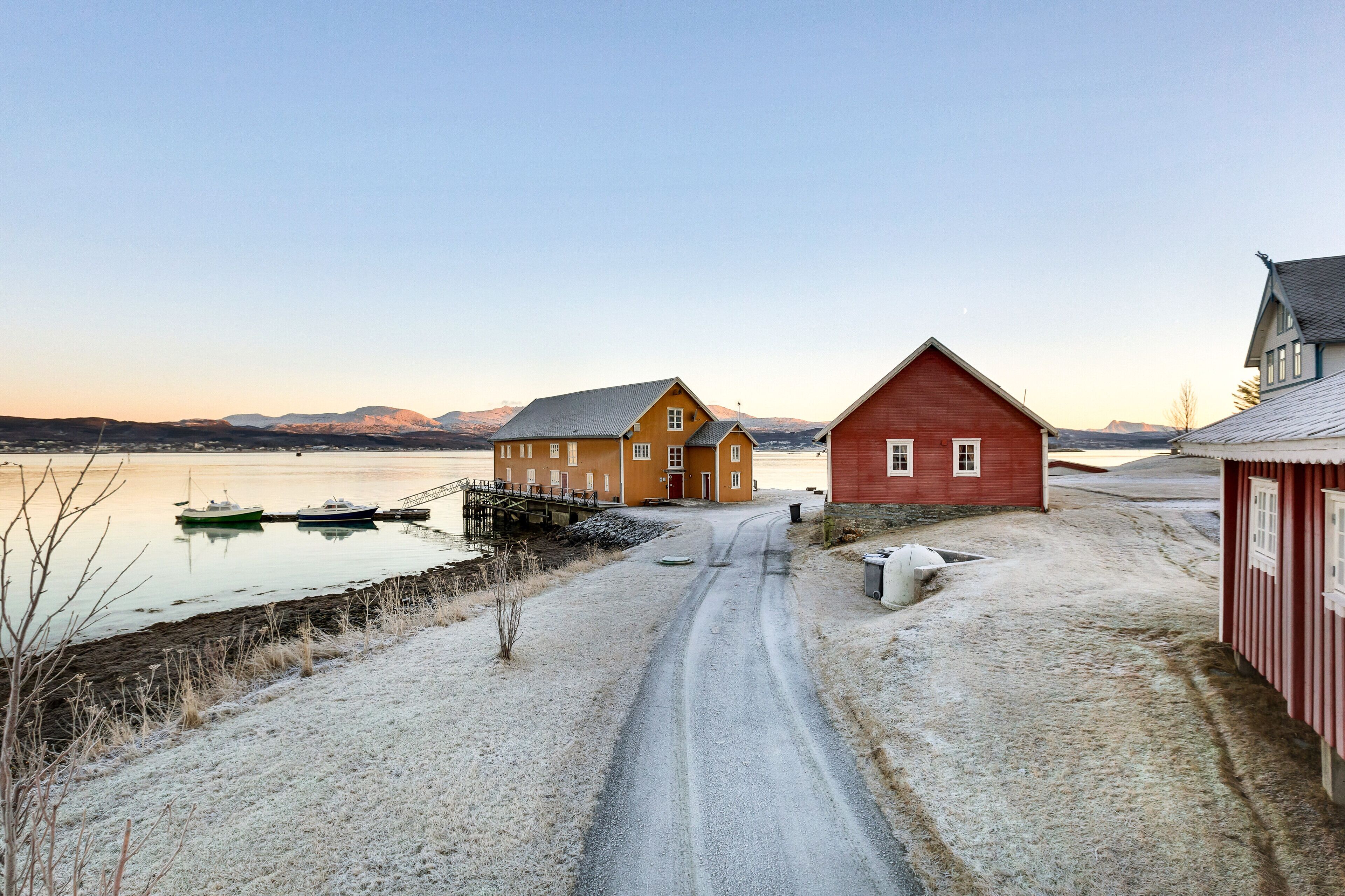 A rural road leading to a yellow house with a red barn nearby, a small dock with boats, and a snowy landscape with mountains in the background.