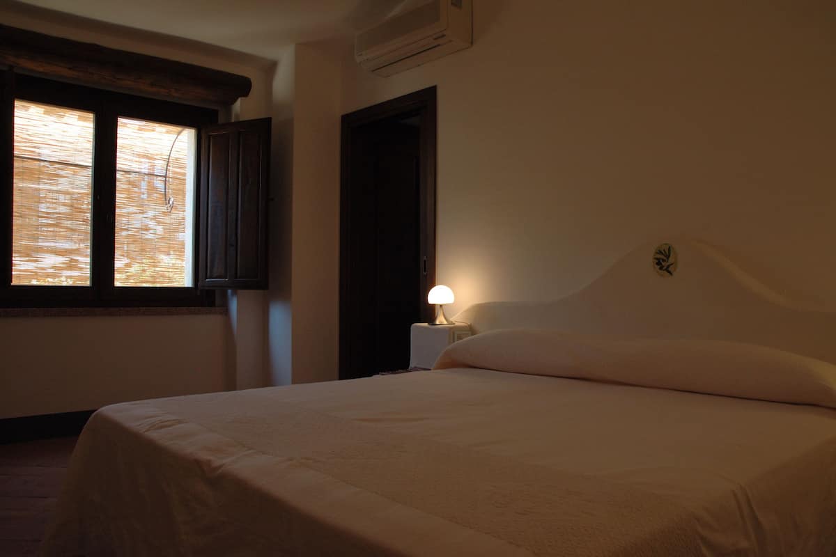 Ein Schlafzimmer mit einem Bett, einem Nachttisch mit Lampe, einem Fenster mit Jalousien und einer Klimaanlage.
