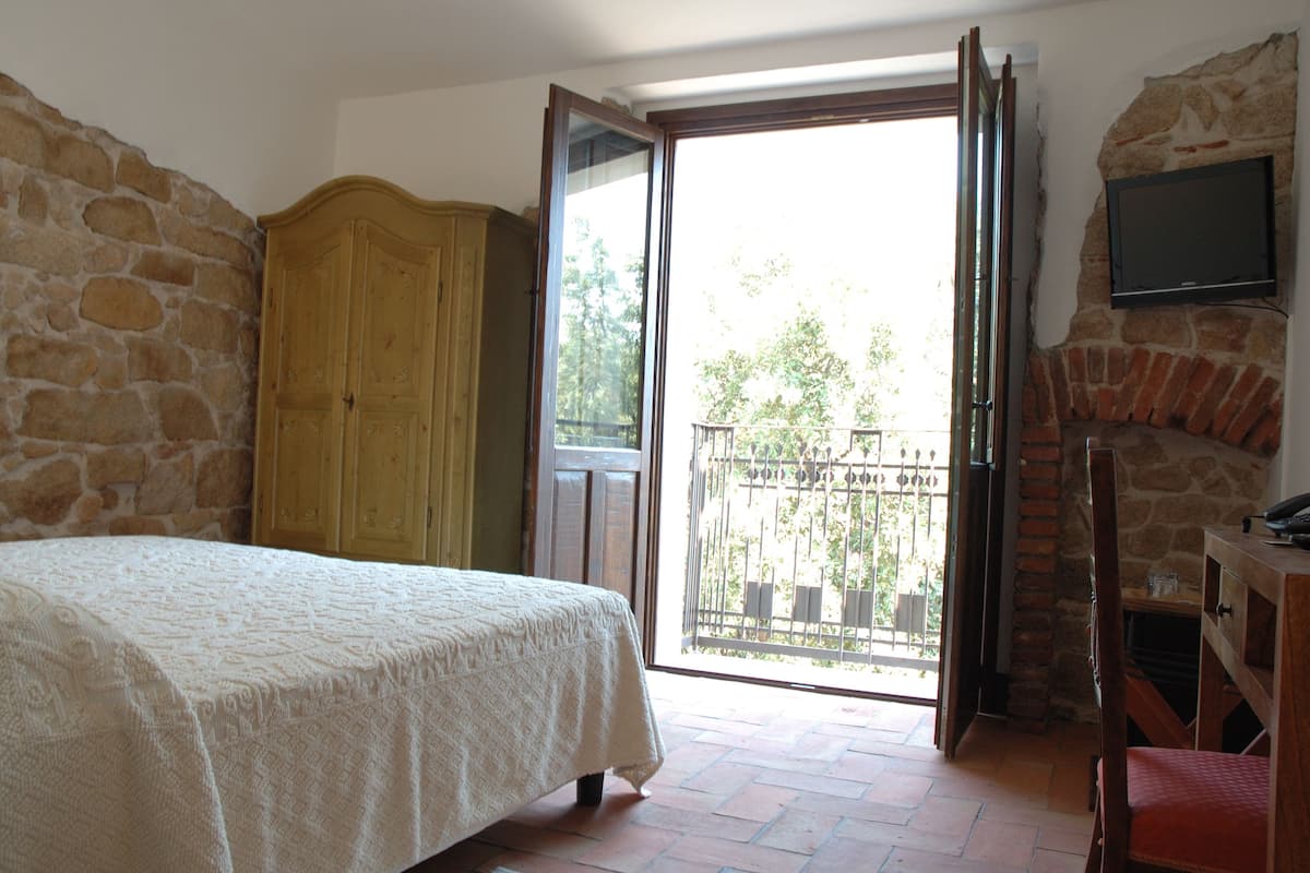 Ein Schlafzimmer mit einem Bett, einem Holzschrank, einem an einer Steinwand montierten Fernseher und einem Balkon mit Blick auf Grünflächen.