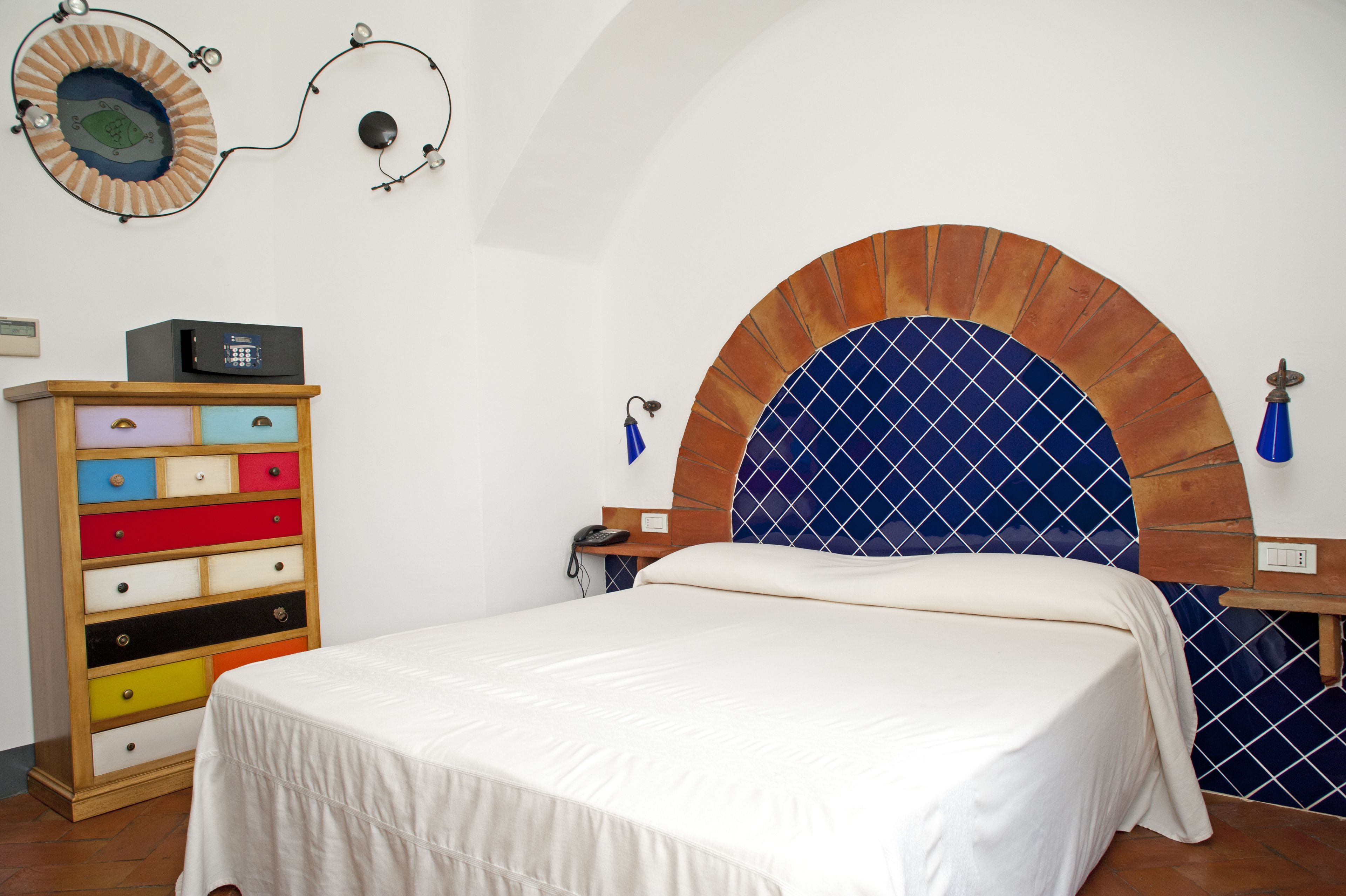 Un dormitorio con cama, cómoda de madera con cajones, cabecera de azulejos azules y lámpara fijada a la pared.