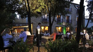 An outdoor dining area with tables and chairs, surrounded by trees and a stone building with balconies. - Nascar Hotel (Baunei)