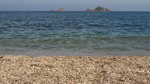 A pebbled beach with clear water and distant rocky islands. - Nascar Hotel (Baunei)