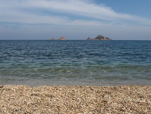 Una playa de guijarros con aguas transparentes y unas islas rocosas a lo lejos.