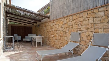 Eine Terrasse im Freien mit Steinmauern, zwei Liegestühlen, einem Tisch mit Stühlen und einer Pergola, die Schatten spendet.