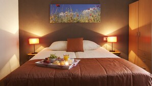 A neatly made bed with a brown blanket, two bedside lamps, a tray with fruits and juice, and a painting of flowers on the wall. - Appartementen Valkenburg (Valkenburg aan de Geul)