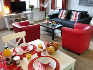Un salon avec des fauteuils rouges, une table blanche dressée pour le petit-déjeuner, un téléviseur et un canapé agrémenté de coussins à rayures.