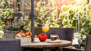 A round wooden table with a wicker basket, croissants, a red bowl, a glass of orange juice, a red mug, and a plate with a coffee cup and a slice of cake. - Appartementen Valkenburg (Valkenburg aan de Geul)