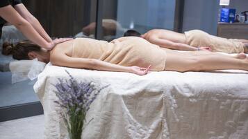 Sala de massagem com dois clientes deitados em macas, um terapeuta atendendo a um cliente, e um vaso de flores no chão.