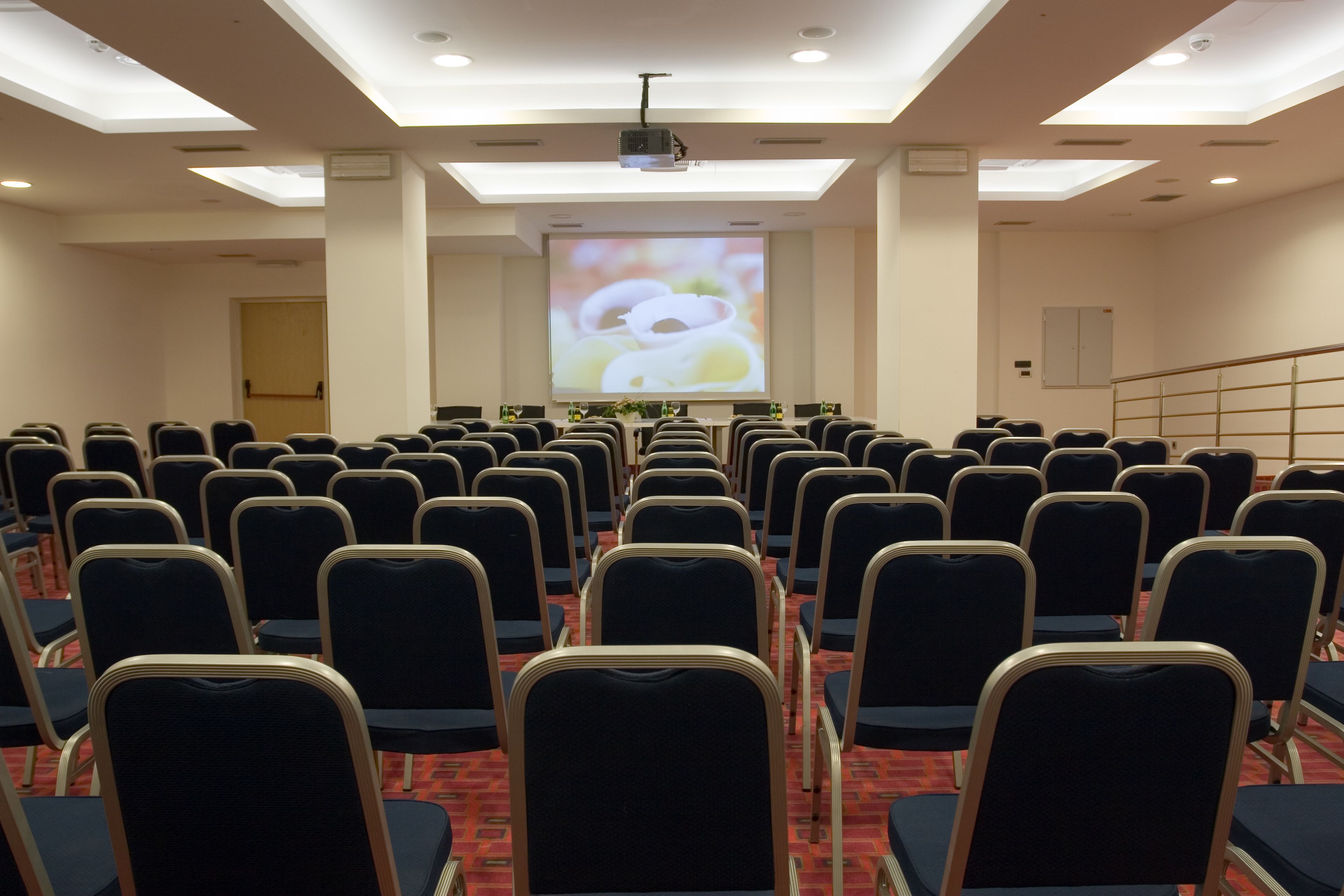 Un salón de conferencias con filas de sillas, una pantalla de proyector y una pantalla grande que muestra una imagen.
