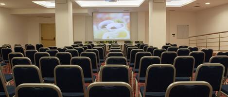Un salón de conferencias con filas de sillas, una pantalla de proyector y una pantalla grande que muestra una imagen.