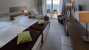 A hotel room with two beds, a sofa, a desk with a lamp, a TV, and a view of the sea.