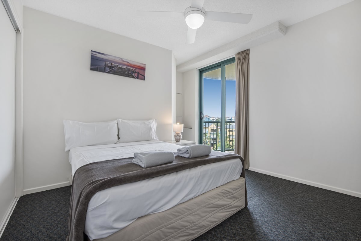 A hotel room with a bed, a ceiling fan, a window with curtains, a painting on the wall, and a view of buildings outside.