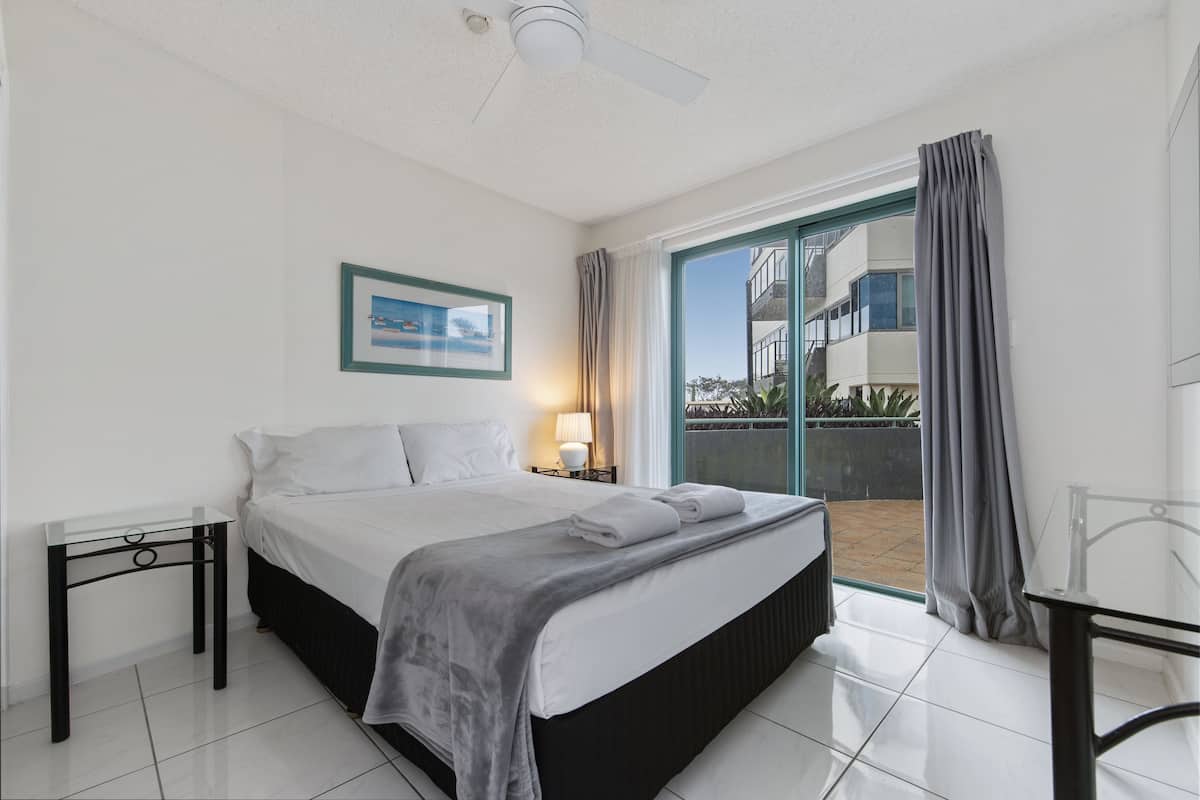 A hotel room with a bed, a nightstand, a ceiling fan, a window with curtains, and a view of the outside.