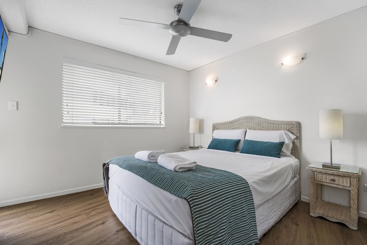 A neatly made bed with a striped bedspread, a ceiling fan, a bedside lamp, and a window with blinds.