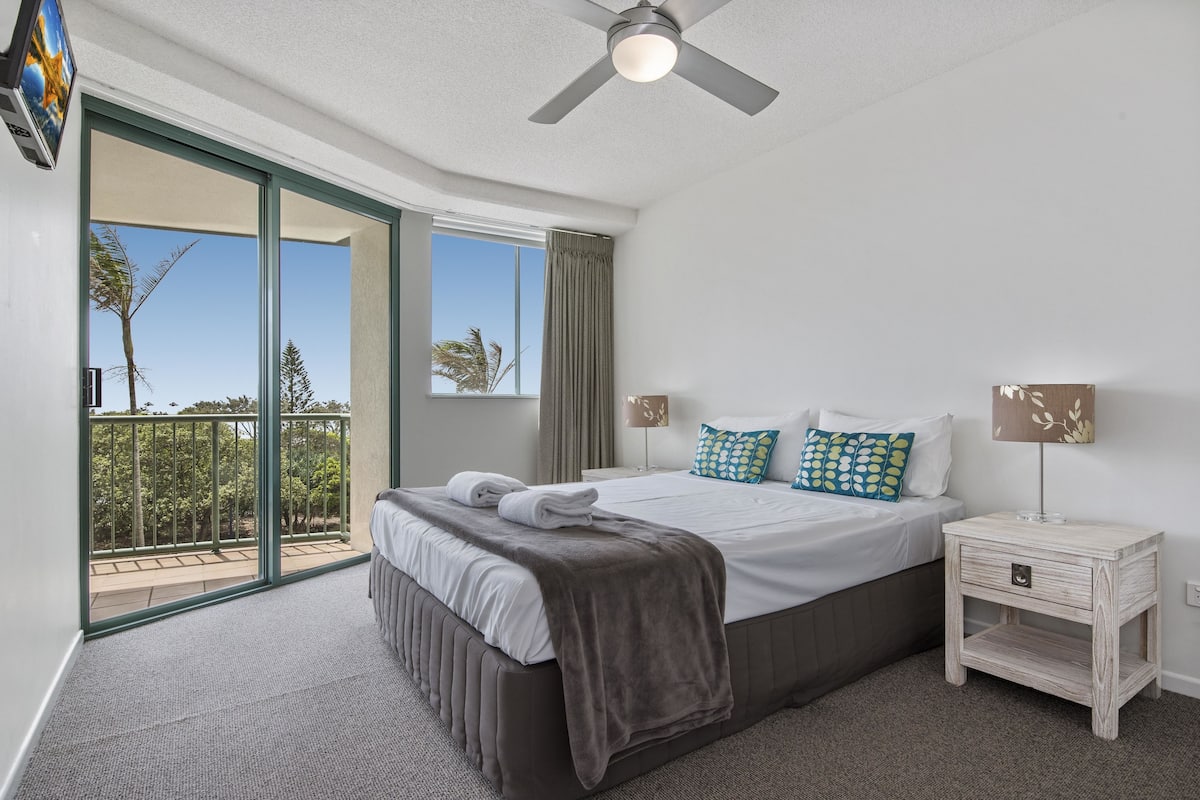 A modern bedroom with a large bed, a nightstand, a ceiling fan, and a balcony with a view of palm trees.