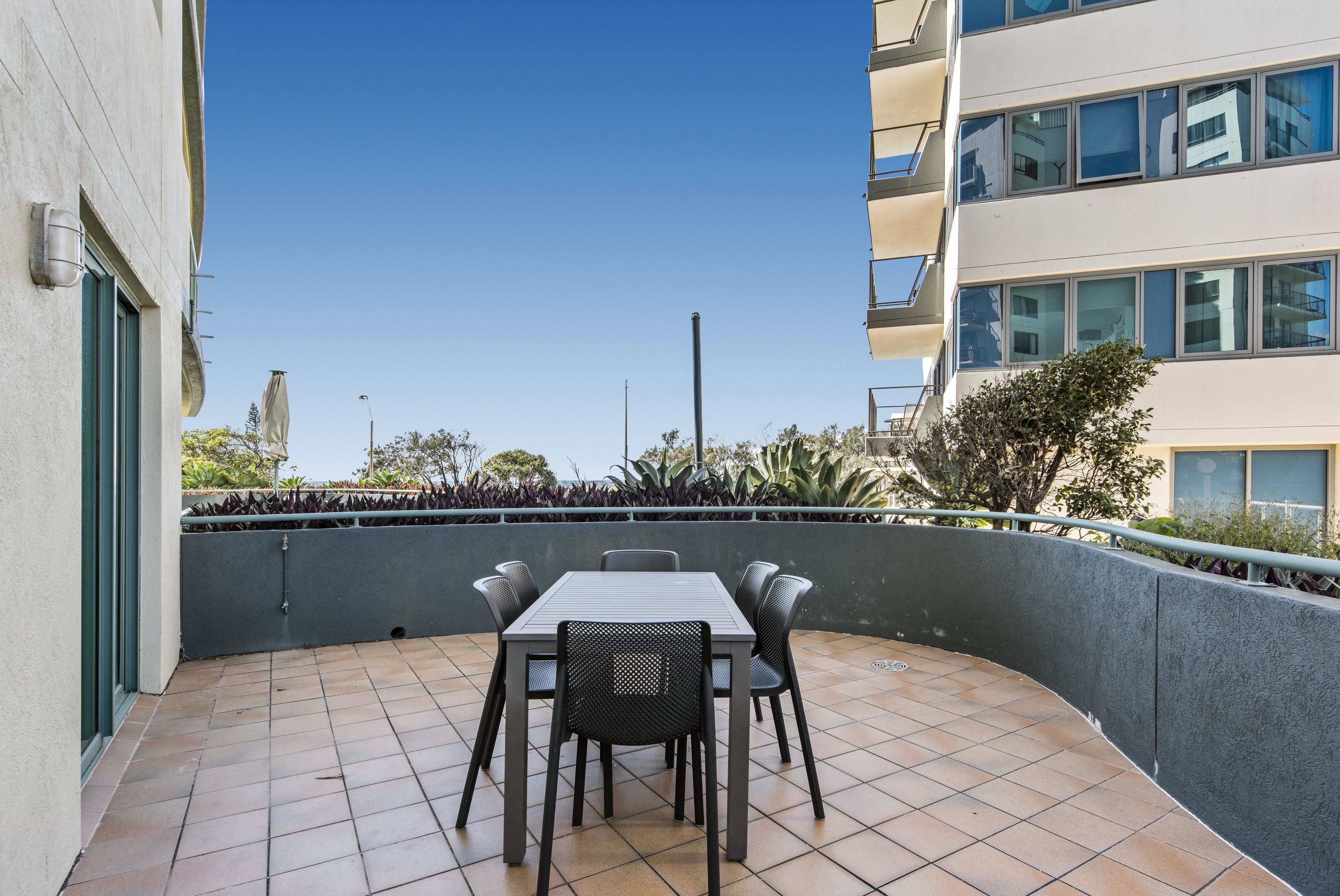 A balcony with a table and chairs, a tiled floor, and a view of buildings and greenery.
