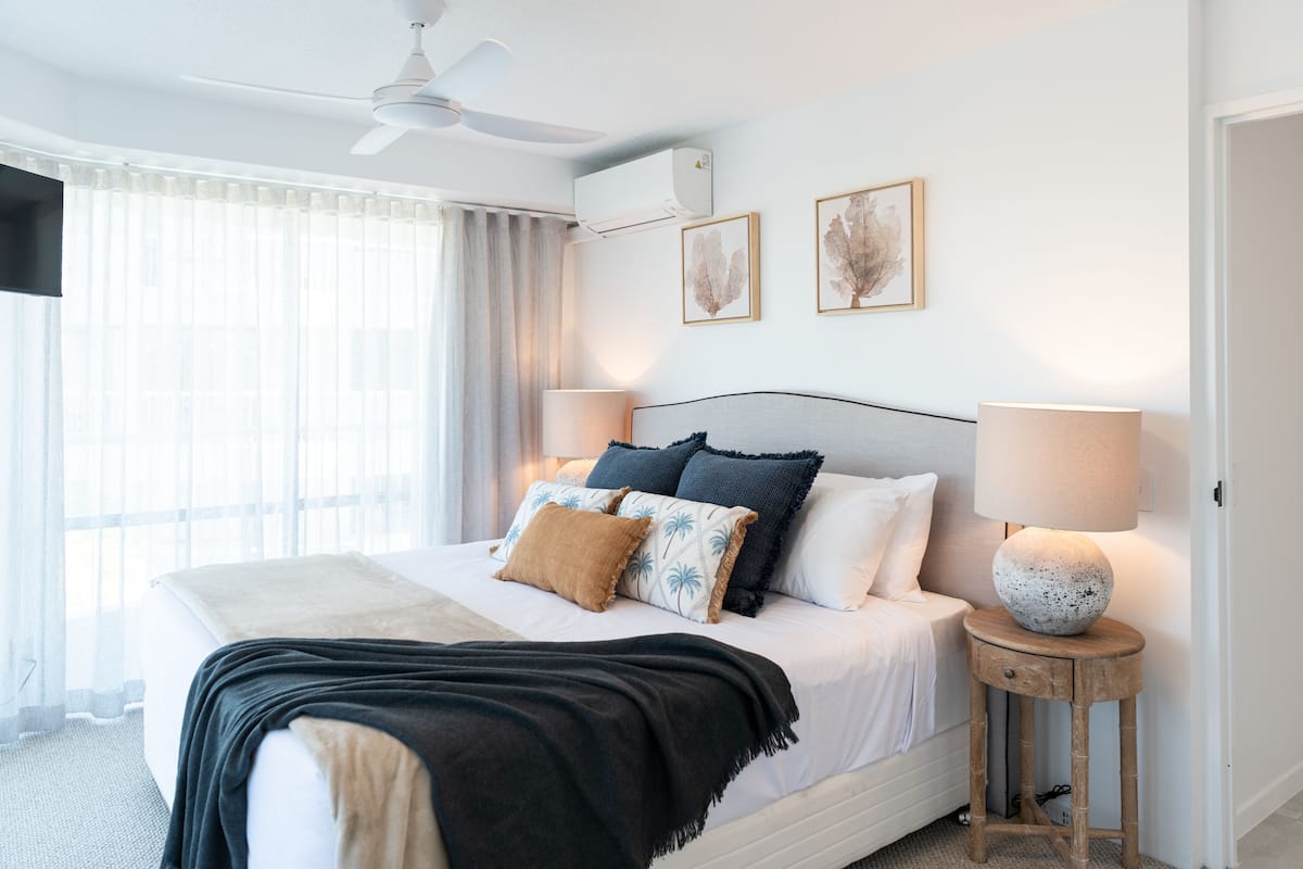 Un dormitorio con una cama, un ventilador de techo, un equipo de aire acondicionado, una lámpara de noche y dos cuadros en la pared.