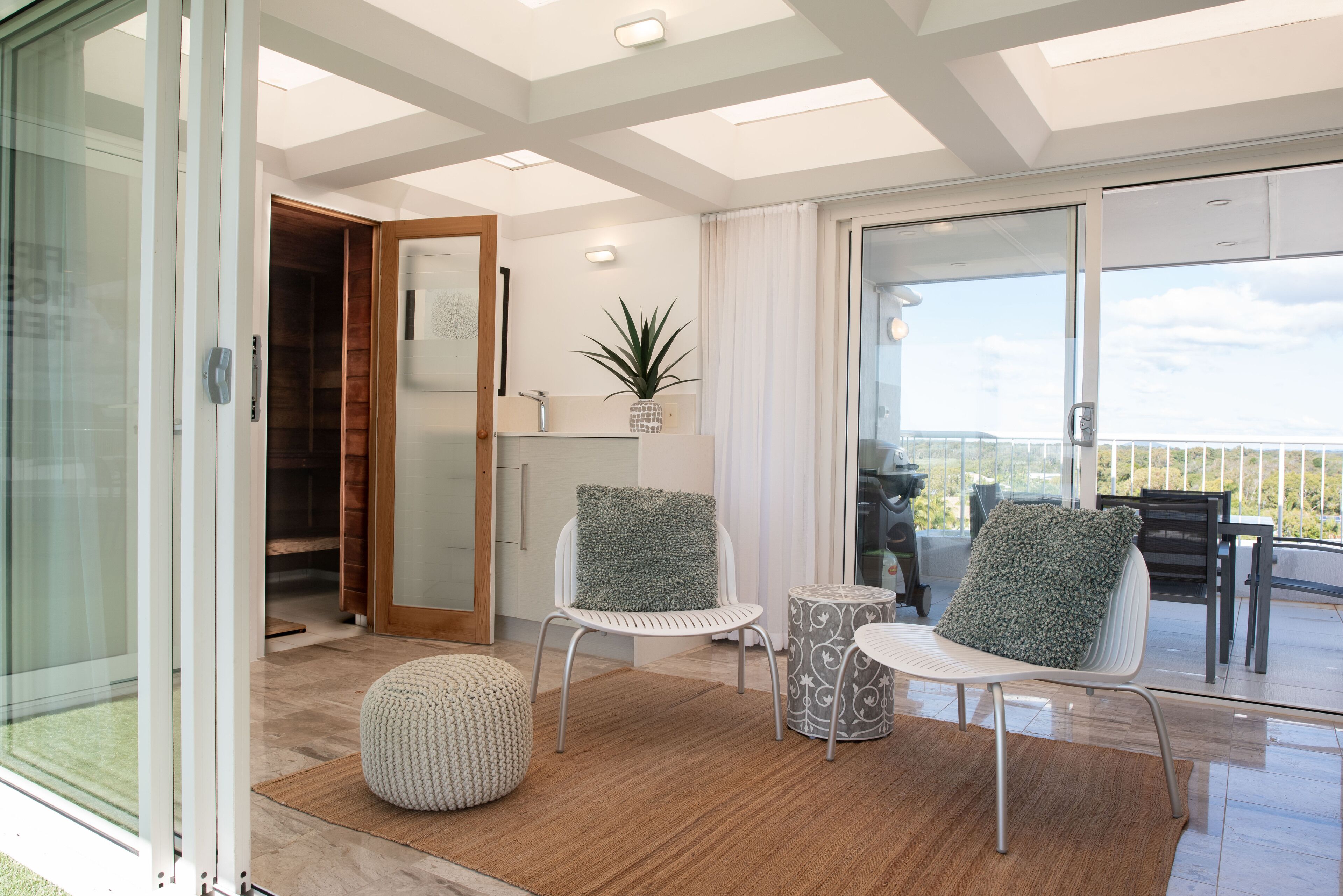 A modern living room with a glass door leading to a balcony, a round ottoman, two white chairs with green cushions, and a small table.