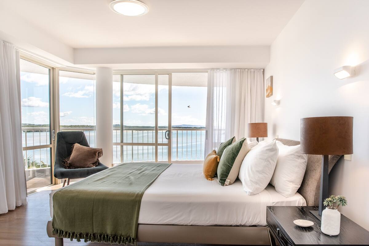 Un dormitorio con una cama grande, una silla, una mesita de noche y vistas al mar.