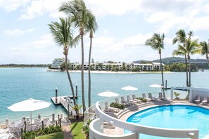 Área de piscina com espreguiçadeiras e guarda-sóis, com vista para um corpo de água e um hotel na praia.