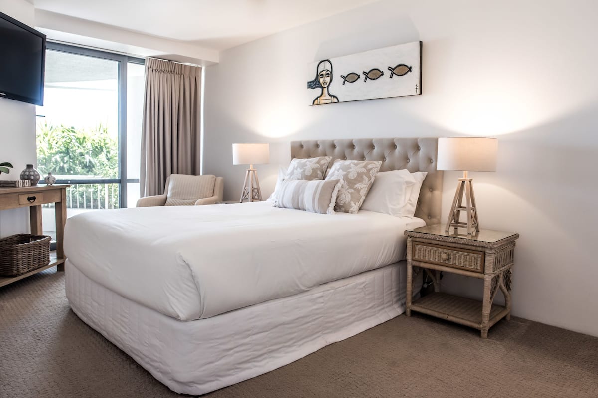 Habitación de hotel con una cama grande, dos mesitas de noche, un escritorio, una silla, un ventanal con cortinas y una obra de arte colgada en la pared.