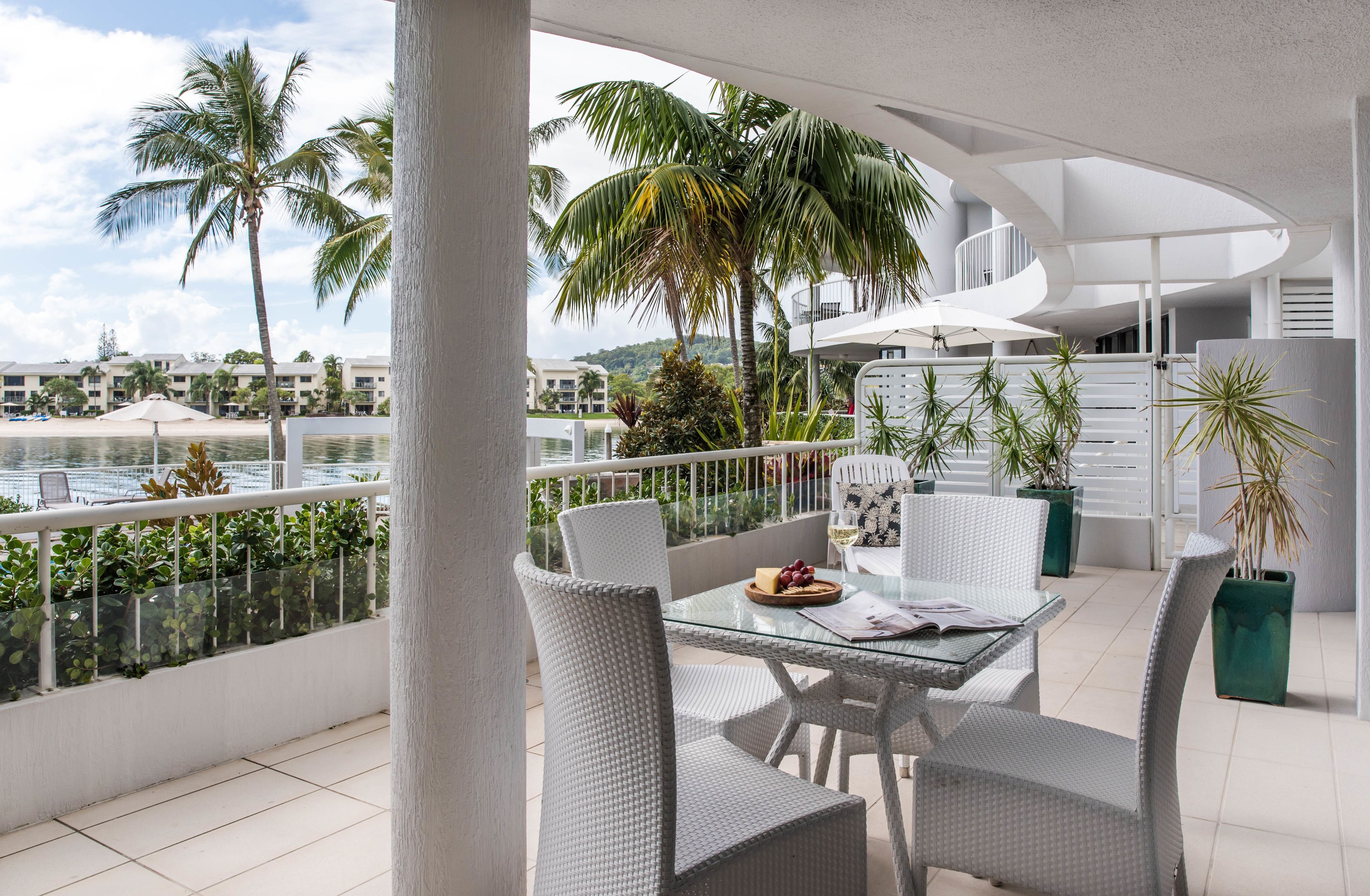 A balcony with a glass table, white wicker chairs, and a view of the beach and palm trees.