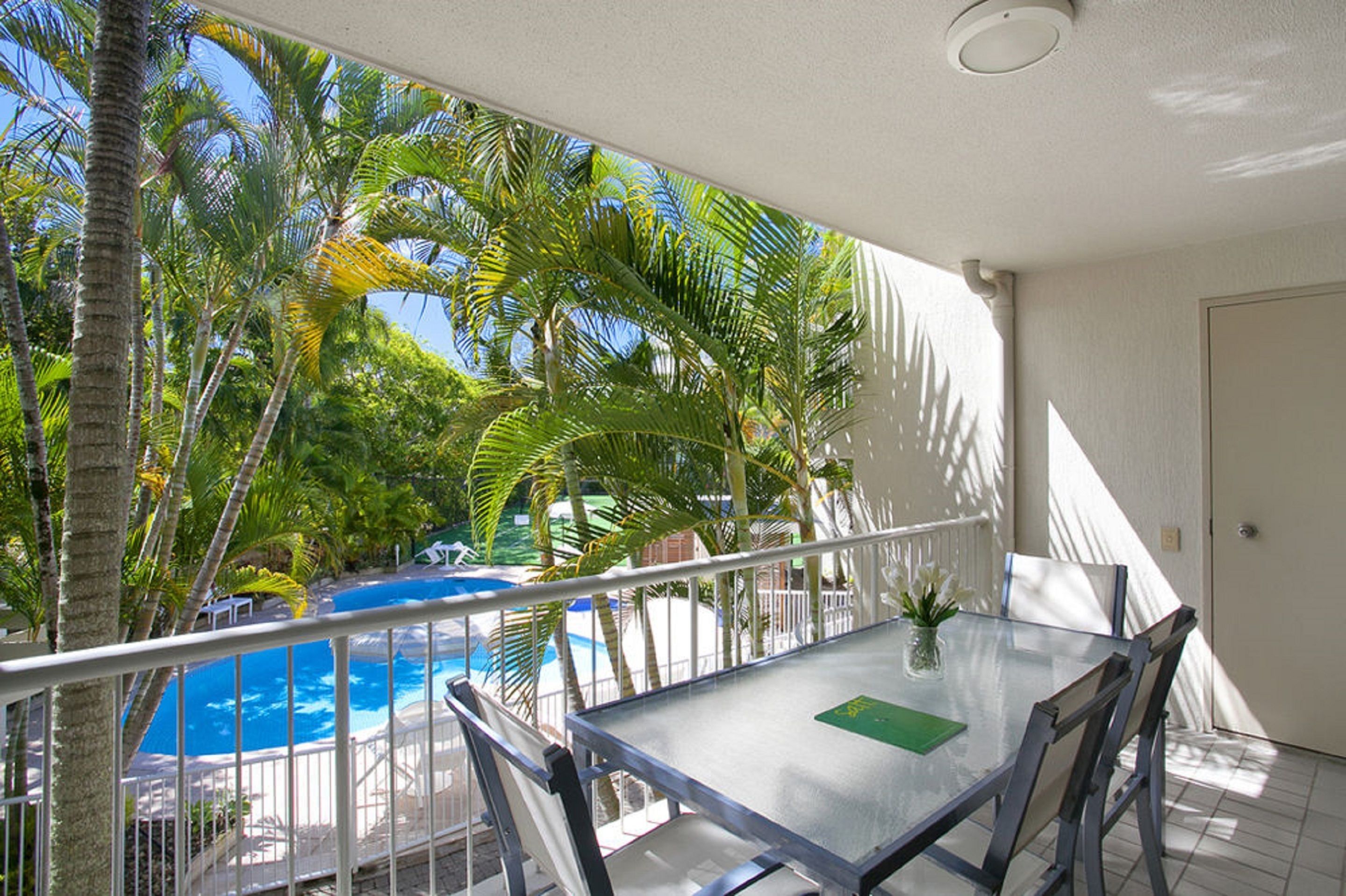 A balcony with a glass table, chairs, and a view of a swimming pool surrounded by palm trees.