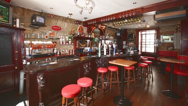 A bar with a stone wall, wooden bar counter, red stools, and a television. - Orchard Hotel (Falkirk)