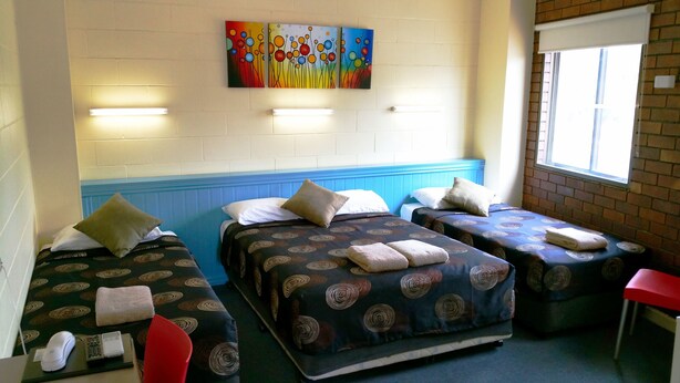 A room with three beds, each with patterned bedding and pillows, a red chair, a small table with a phone and remote, and a window with a view of the outside. - A & A Lodge Motel Emerald (Emerald)