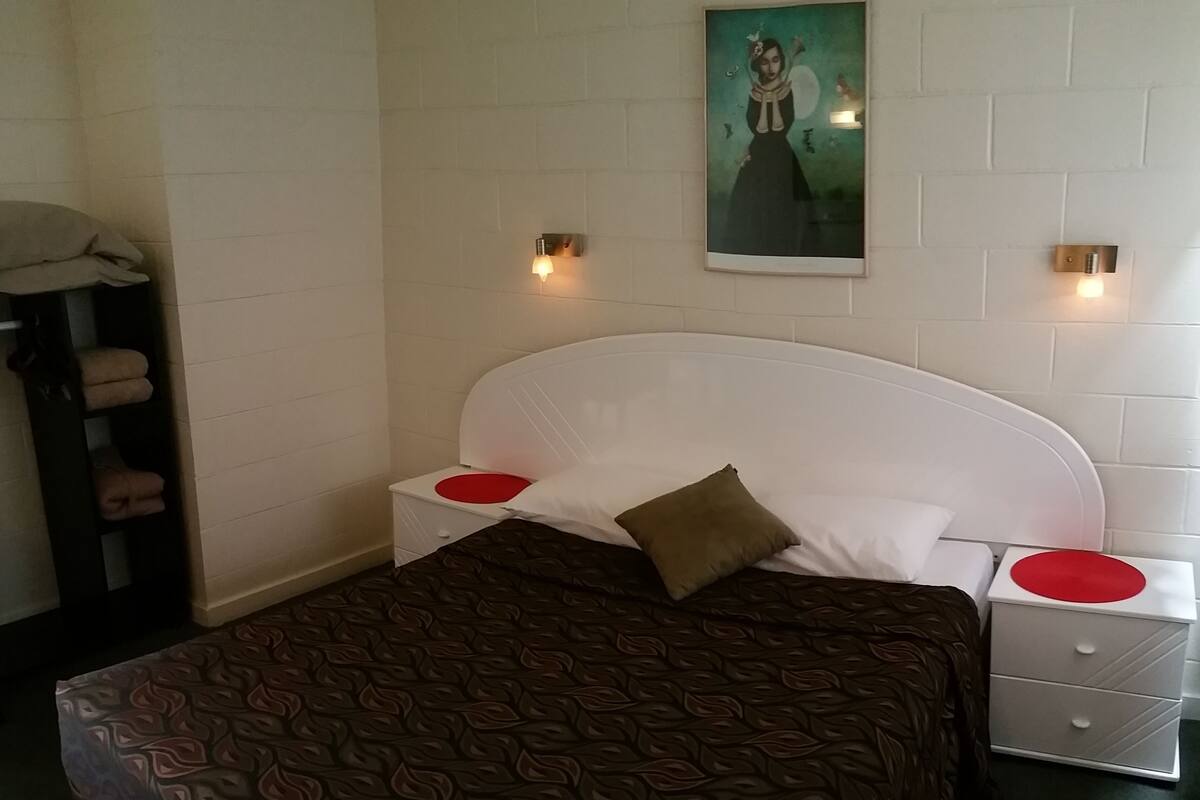 Habitación de hotel con una cama, dos mesitas de noche, una lámpara de noche, un cuadro colgado en la pared y un estante con toallas.