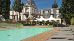 A large pool with a wooden deck, surrounded by lounge chairs and umbrellas, in front of a historic building with multiple windows and a balcony. - LE H - Hôtel & Restaurant (Barr)