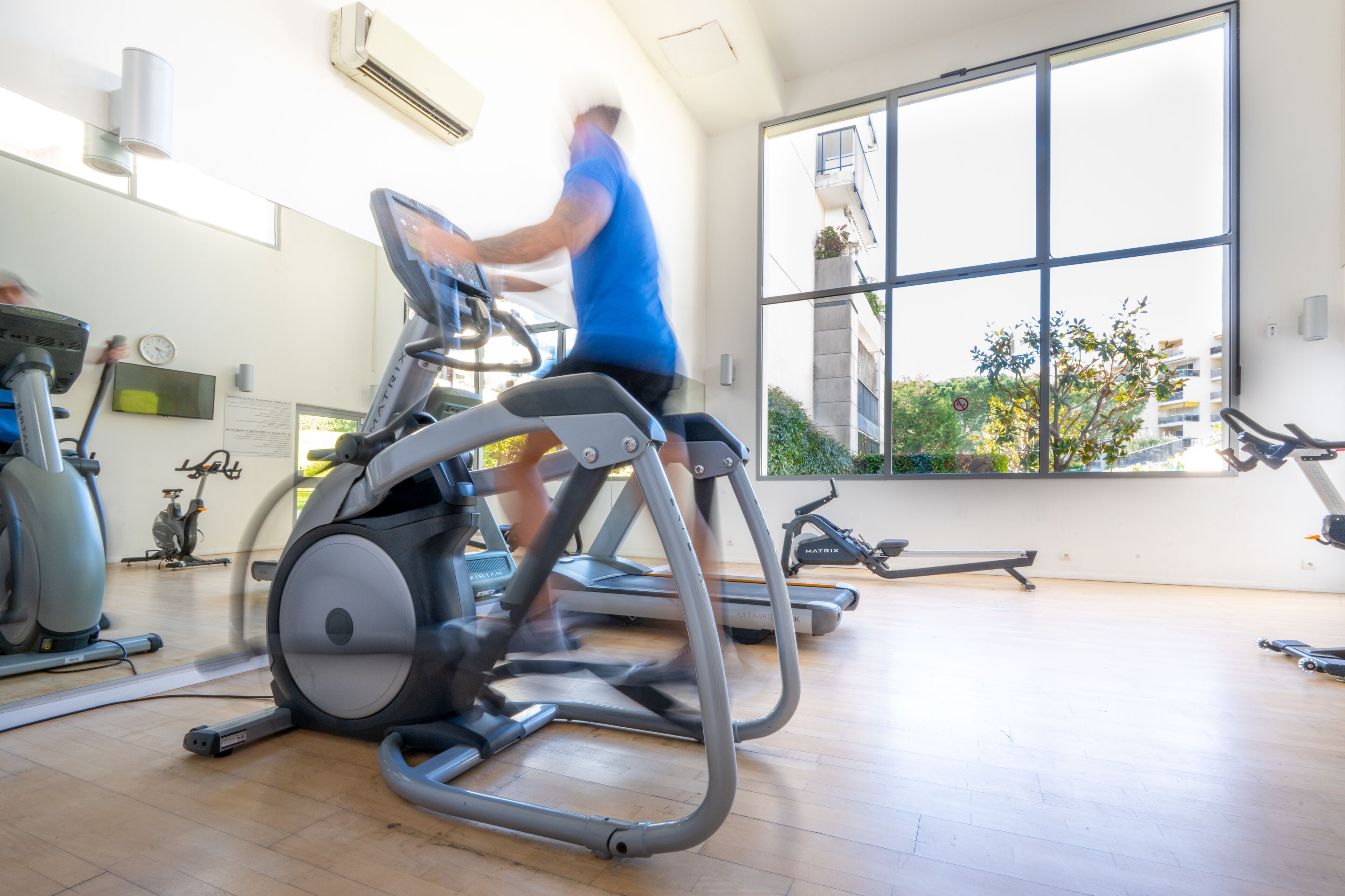 A gym with stationary bikes, treadmills, and a large window offering a view of buildings and greenery.