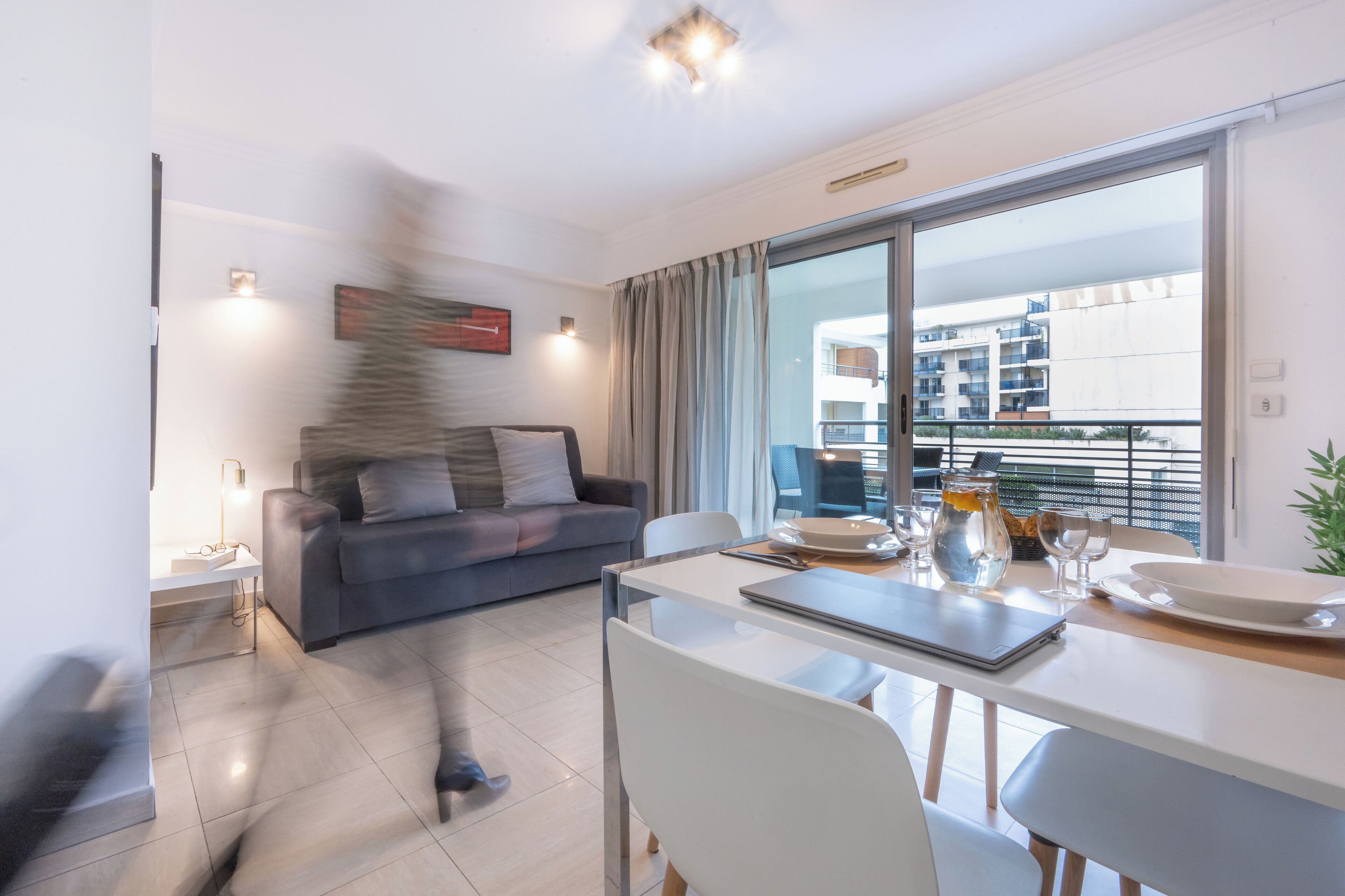 Una sala de estar moderna con un sofá gris, una mesa de comedor con sillas y un ventanal con vista a un edificio.