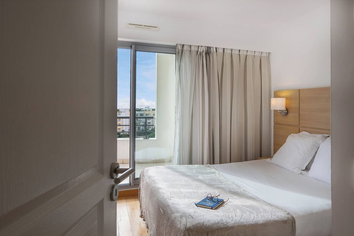 A hotel room with a bed, a nightstand, a lamp, and a view of the city through the window.